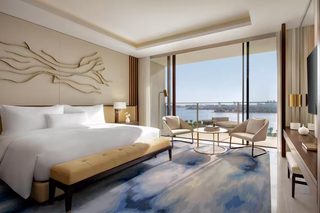A modern hotel room with a large bed, a bench, a round table, two chairs, a TV, and a view of the city.
