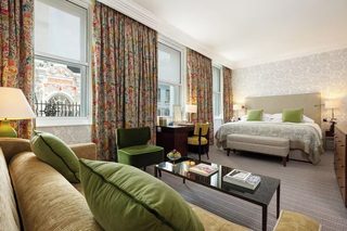 A hotel room with a bed, a green armchair, a desk, a coffee table, and a view of a building outside.