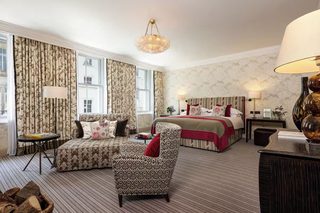 A spacious bedroom with a large bed, a sitting area with a patterned armchair, and a desk with a lamp.