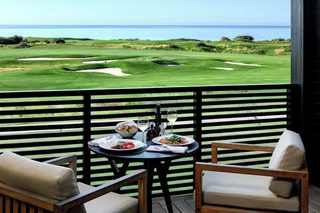 A dining table with plates of food and drinks, overlooking a golf course and the ocean.