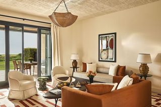A living room with a sofa, chairs, a coffee table, a hanging light fixture, a painting, and a view through a glass door.