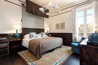 A hotel room with a large bed, two bedside lamps, a decorative chandelier, a view of the city through the window, and a blue armchair.