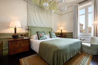 A hotel room with a large bed, two bedside tables, a chair, a window with a view, and a chandelier.