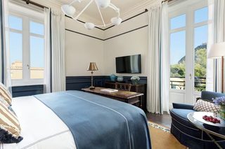 A hotel room with a large bed, a desk with a lamp, a TV, and a balcony with a view of the city.