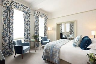 A hotel room with a large bed, two armchairs, a small table with a lamp, and floral curtains.
