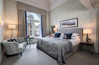 A hotel room with a large bed, a sofa, a small table, and a window with a view of buildings.