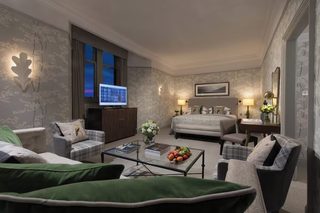A hotel room with a bed, two armchairs, a sofa, a coffee table with books and fruit, a TV, and a cityscape view.