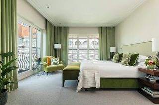 A hotel room with a large bed, a green armchair, a small table with a lamp, and a view of buildings through large windows.