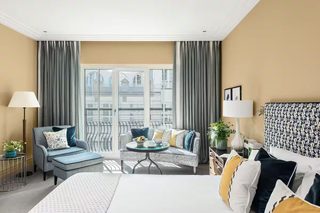 A hotel room with a bed, a sofa, a small round table with a bowl of fruit, and a view of a building through large windows.