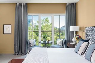 A hotel room with a large bed, two armchairs, a small table with a vase of flowers, and a view of a balcony with greenery and buildings.