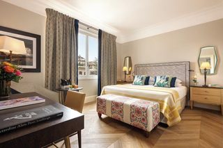 A hotel room with a bed, bedside tables, a desk, a chair, a window with curtains, and a view of buildings outside.