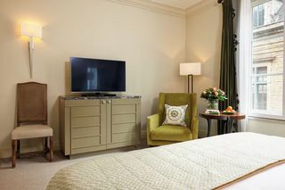 A hotel room with a bed, a television on a cabinet, a chair, a small table with a vase and fruit, and a window with curtains.