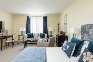 A hotel room with a large bed, a sofa, a desk, and a chair. There are two lamps, a window with curtains, and a framed picture on the wall.
