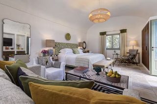 A well-lit bedroom with a bed, bedside tables, a chair, a small table, and a window with curtains.