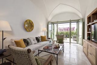 A modern living room with a sofa, coffee table, and wicker chairs. A large window offers a view of the outdoors.