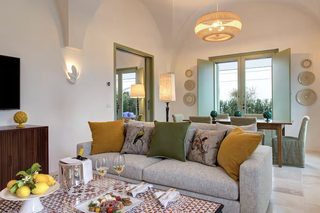 A modern living room with a sofa, dining table, and chairs. There is a television on the wall and a window with a view of greenery.