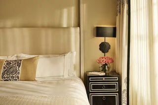 A neatly made bed with white linens and decorative pillows, a bedside table with a lamp and a vase of flowers, and a window with sheer curtains.