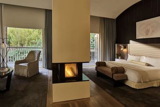 A modern hotel room with a fireplace, a balcony with a view of greenery, a sofa, and a bed with a headboard.