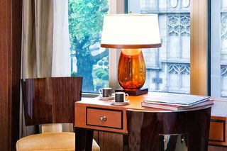 A hotel room with a wooden desk, a lamp, a chair, and a view of a building through a window.