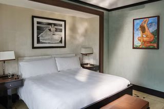 A hotel room with a large bed, a bedside table with a lamp, a bench, and two framed artworks on the walls.