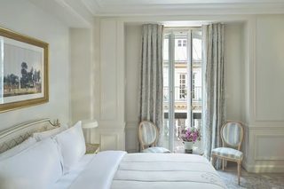 A hotel room with a bed, two chairs, a table with a vase of flowers, and a balcony with curtains.