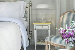 A neatly arranged bedroom with a bed, a bedside table with a lamp, a chair, and a framed picture on the wall.
