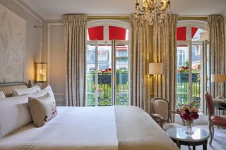 A hotel room with a large bed, a sitting area with a table, and a view of a balcony with red awnings.