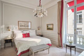 A hotel room with a large bed, a bench, a chandelier, a window with red curtains, and two paintings on the wall.