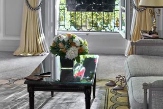 A room with a large window, a table with a vase of flowers, a chair, and a lamp.