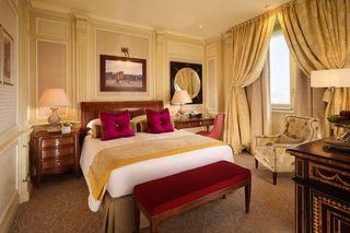 A hotel room with a large bed, a wooden nightstand, a desk, a chair, and a window with curtains.