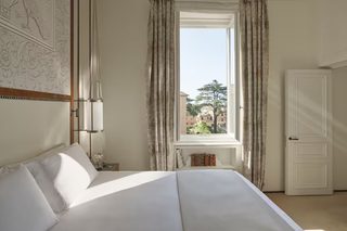 A bedroom with a large bed, a window with curtains, a bedside table with books, and a door.