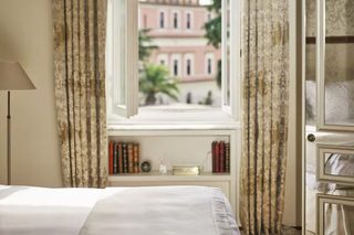 A bedroom with a large window, curtains, a bookshelf, and a lamp.