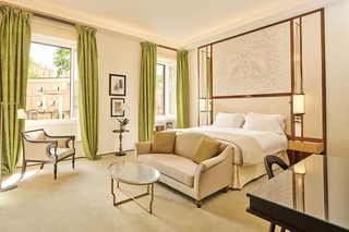A hotel room with a bed, a sofa, a small table, and a chair. There is a window with curtains, a lamp, and a picture on the wall.