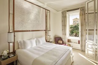A hotel room with a large bed, a nightstand with a lamp, a chair, a window with curtains, and a view of the outside.