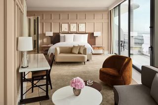 A hotel room with a large bed, a sofa, a chair, a small table, and a view of the city through large windows.