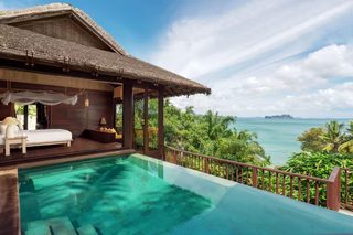 A poolside area with a wooden deck, a lounge chair, and a view of the ocean and palm trees.