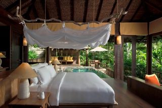 A bedroom with a large bed, a bedside table with a lamp, a view of a pool, and a balcony with outdoor furniture.