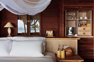 A wooden cabin room with a bed, a nightstand with bottles, a lamp, and a shelf with glasses and a coffee maker.
