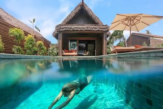 A swimming pool with a person diving, a thatched-roof structure, and lounge chairs.