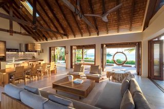 A spacious living area with a high ceiling, wooden beams, and a view of the ocean. There are multiple seating areas, including a sofa and chairs, and a dining table.