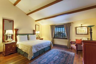 A well-lit bedroom with a large bed, a nightstand, a chair, a window with curtains, and a framed picture on the wall.