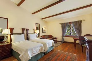 A hotel room with two beds, a desk, a chair, a window with curtains, a rug, and a nightstand with a lamp.