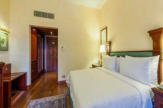 A hotel room with a large bed, a nightstand with a lamp, a wooden desk, and a door leading to another room.