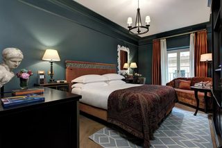 A hotel room with a large bed, a desk with books, a bust sculpture, and a view of the city through the window.