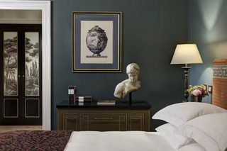 A bedroom with a bed, a dresser with a bust sculpture, a framed artwork, and a nightstand with a lamp and flowers.