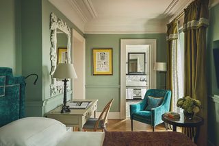 A hotel room with a bed, a desk with a lamp, a chair, a small table with a vase, and a large mirror.