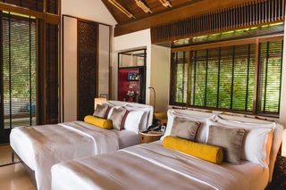 A hotel room with two beds, a minibar, and large windows with blinds.