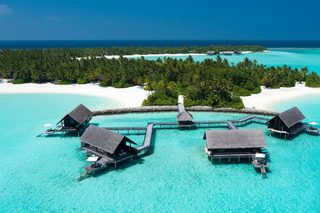An aerial view of overwater bungalows connected by a wooden walkway over turquoise waters, surrounded by lush greenery and white sand beaches.