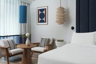 A hotel room with a bed, two armchairs, a round wooden table, a vase with flowers, a framed abstract painting, a woven hanging light fixture, and a side table with books.