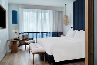 A modern hotel room with a large bed, a desk with a chair, a small table, and a large window with curtains.
